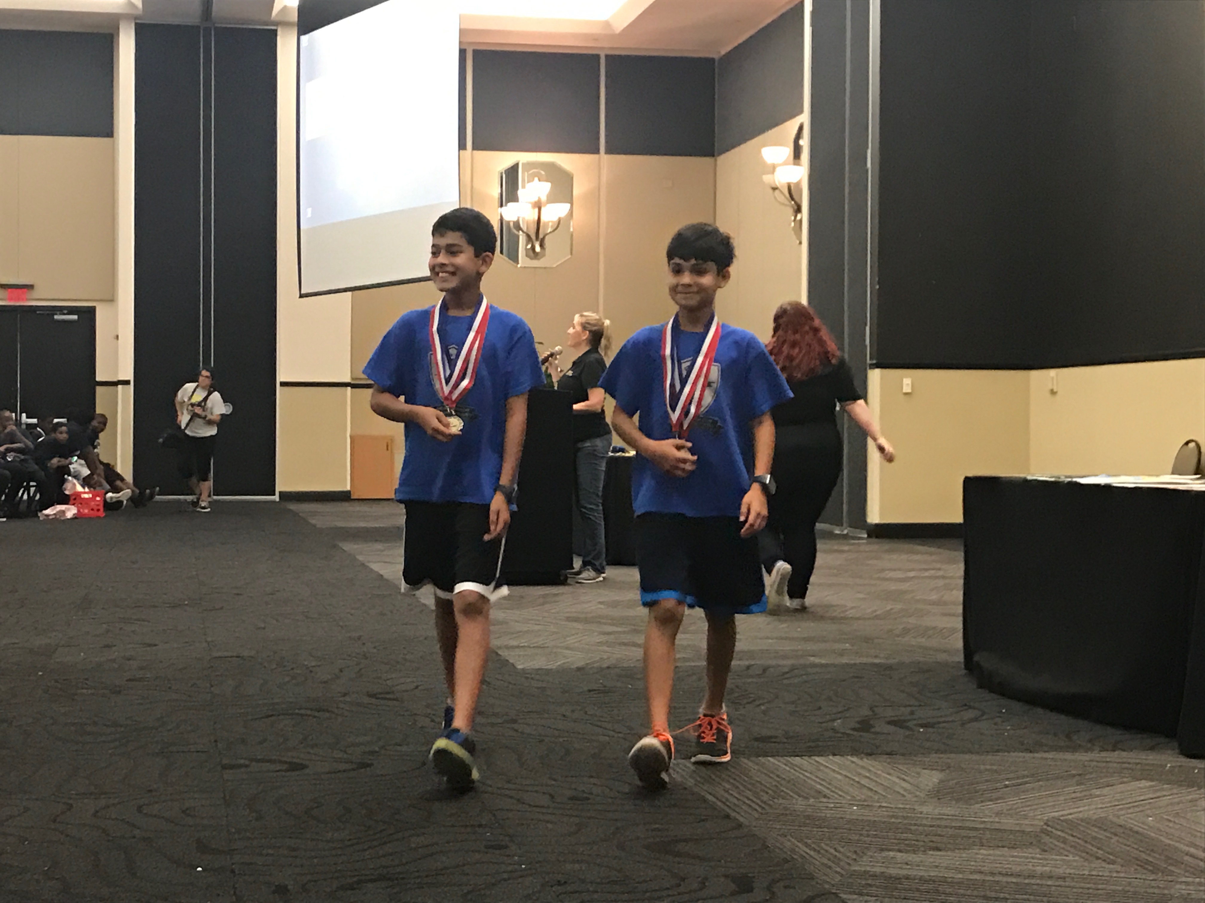 Two boys in matching blue shirts and blue pants holding medals around their necks walking towards the camera