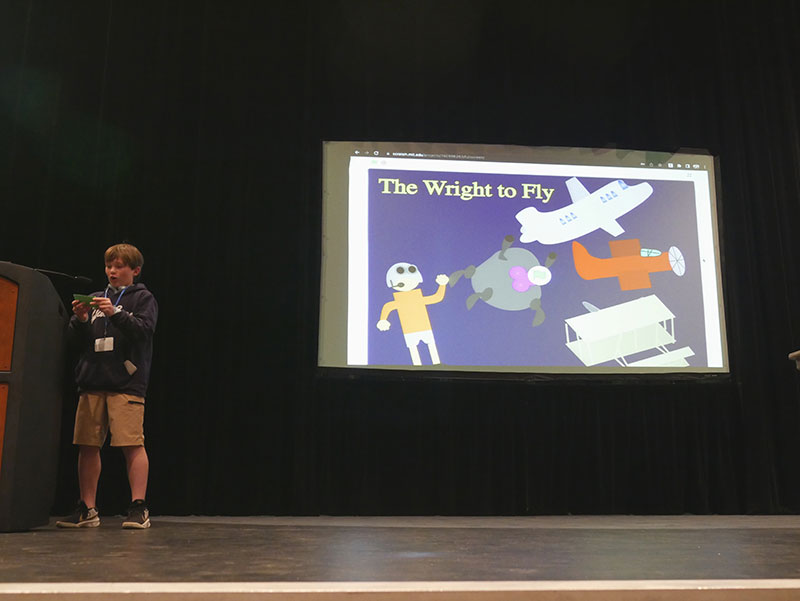 A boy standing on a stage, reading notecards, and speaking into a podium’s microphone. There is a projector screen pictured behind and to the right of him. It is displaying an illustrated person and several planes, with the title “The Wright to Fly” across the top.