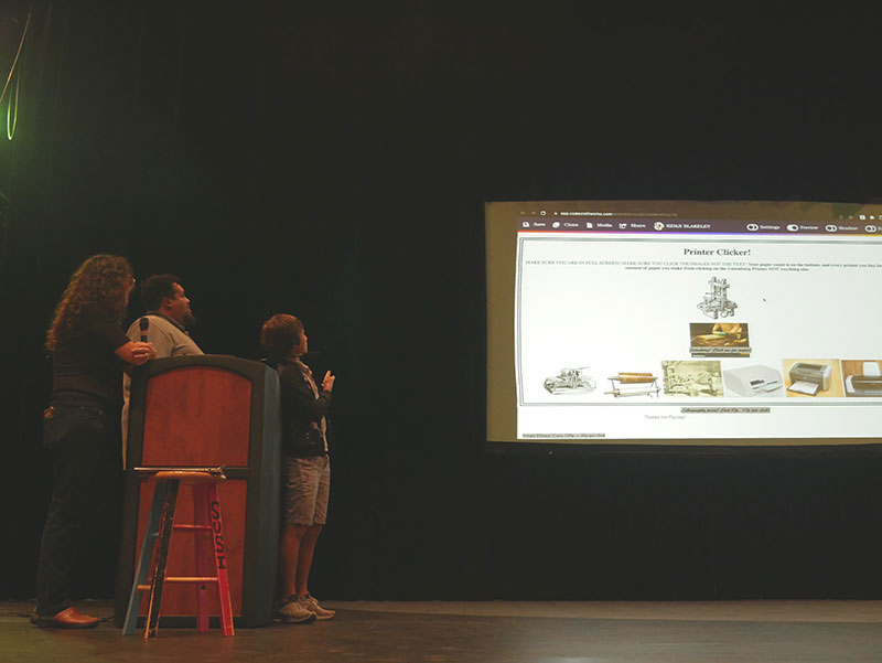 Two adults and a student standing around a podium on a stage, all facing a projector screen that is pictured behind and to the right of them. The projector displays a webpage titled "Printer Clicker!" and features other text and images of various types of printers.