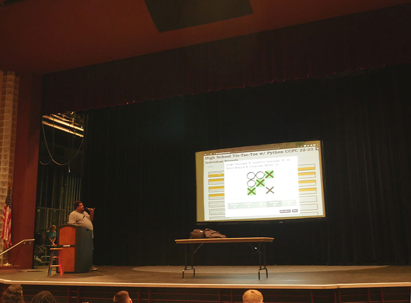 A photo of a man standing in front of a podium onstage in an auditorium, speaking into a microphone and facing a projector screen. The screen shows a completed game of tic-tac-toe, with a diagonal line of "X" marks on the board highlighted in green.