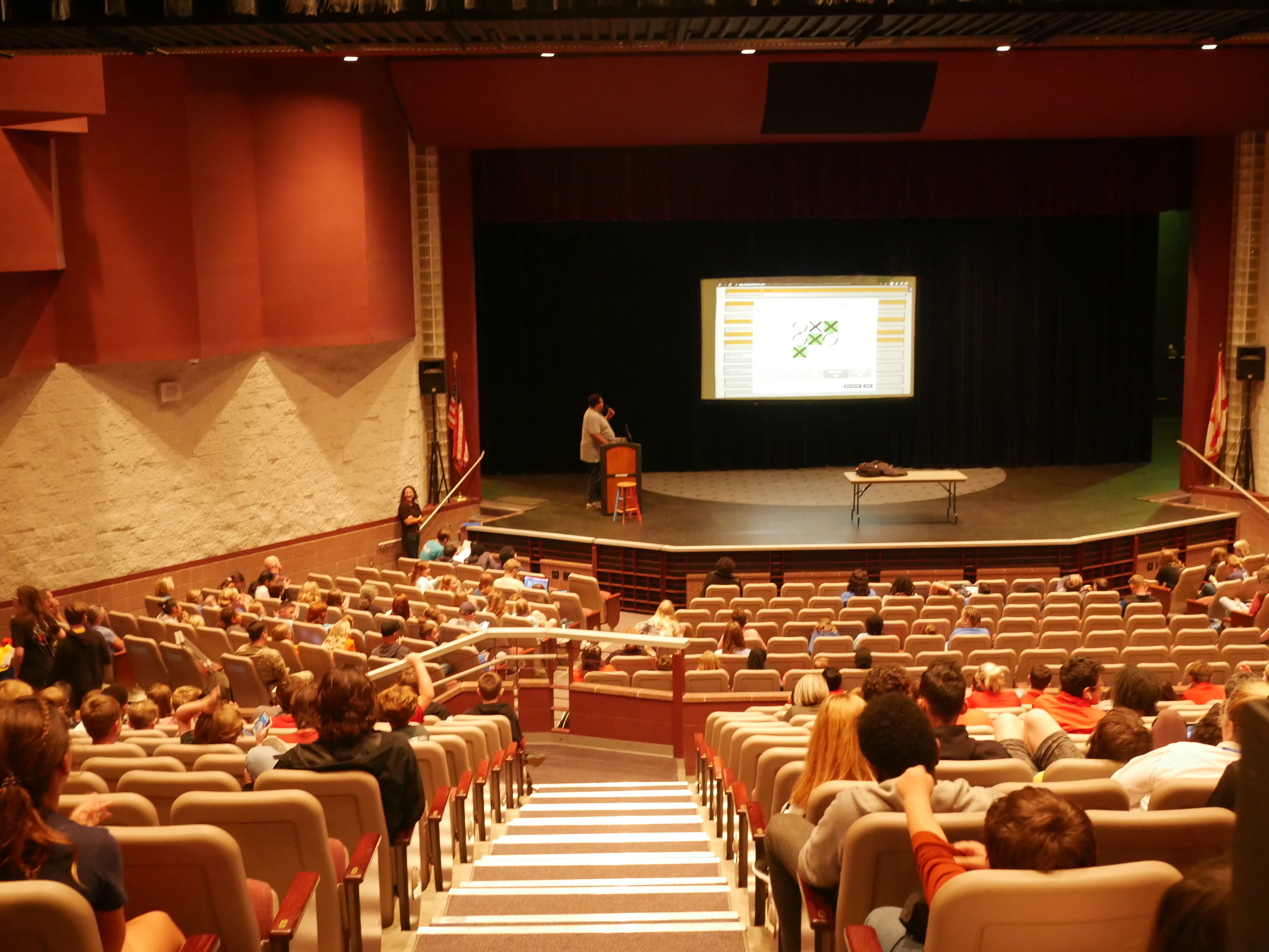 A photo taken from a distance of a man standing in front of a podium onstage in an auditorium, speaking into a microphone and facing a projector screen. Students and teachers are seated in the audience, watching the presenter. The projector screen shows a completed game of tic-tac-toe.