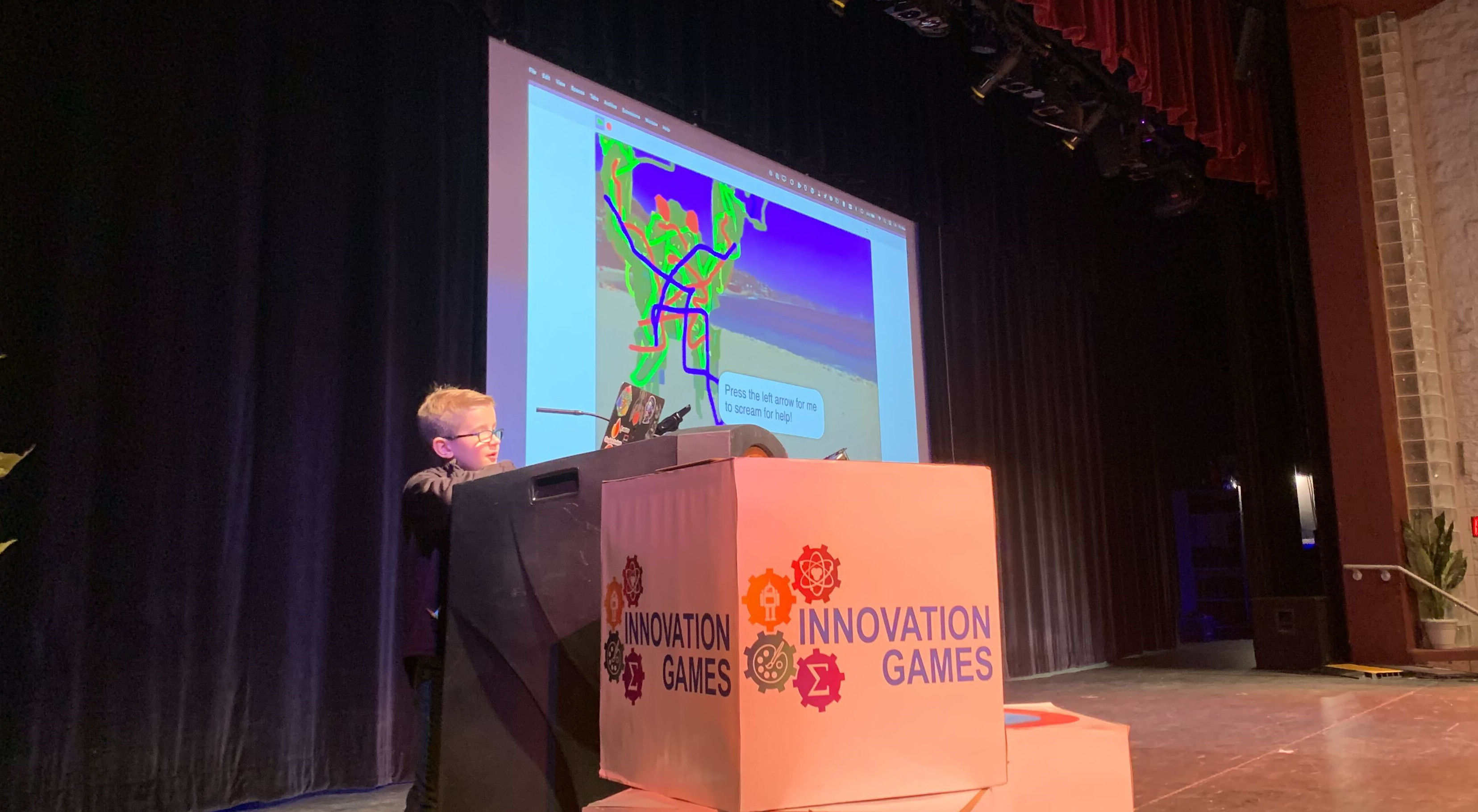 A photo of a boy standing on stage at a podium, looking at a laptop in front of him. A projector screen behind him shows a monster drawn in red, blue, and green squiggly lines and a text bubble that says "Press the left arrow for me to scream for help!"