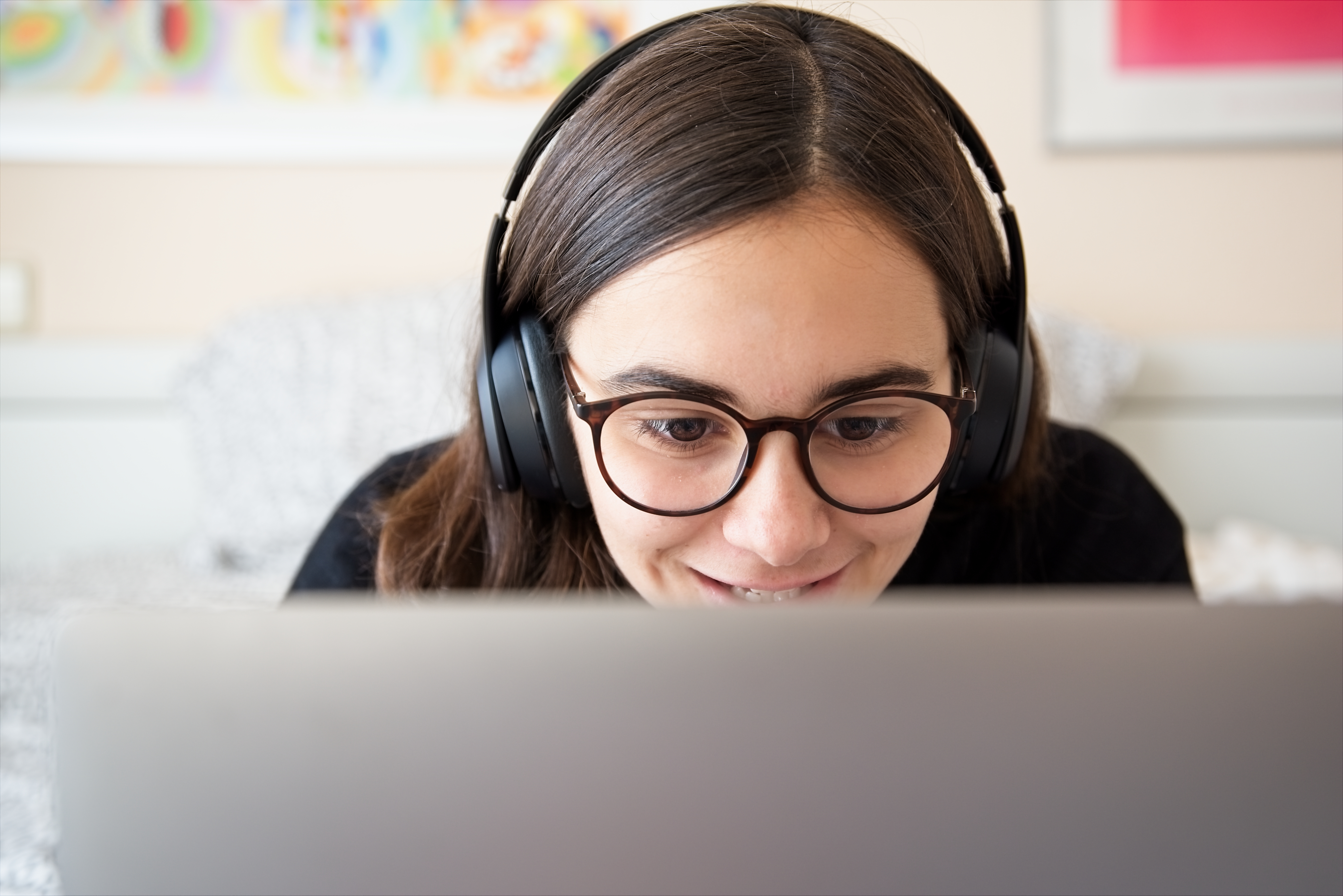 A woman wearing glasses and headphones staring at a computer screen in front of them while smiling